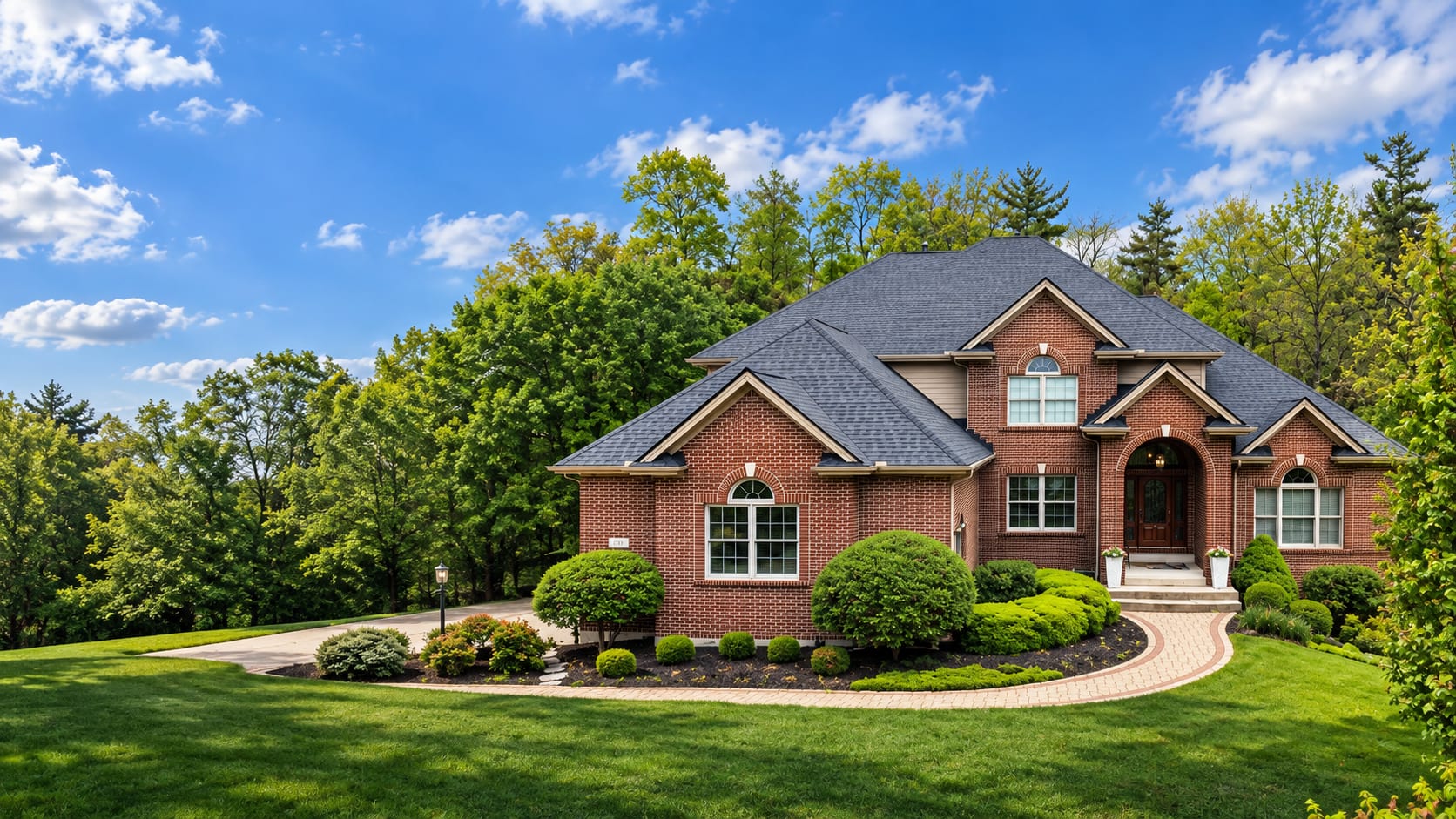 A brick estate under a blue sky with a charcoal architectural shingle roof, trimmed shrubs along the front and a paver walk to the door